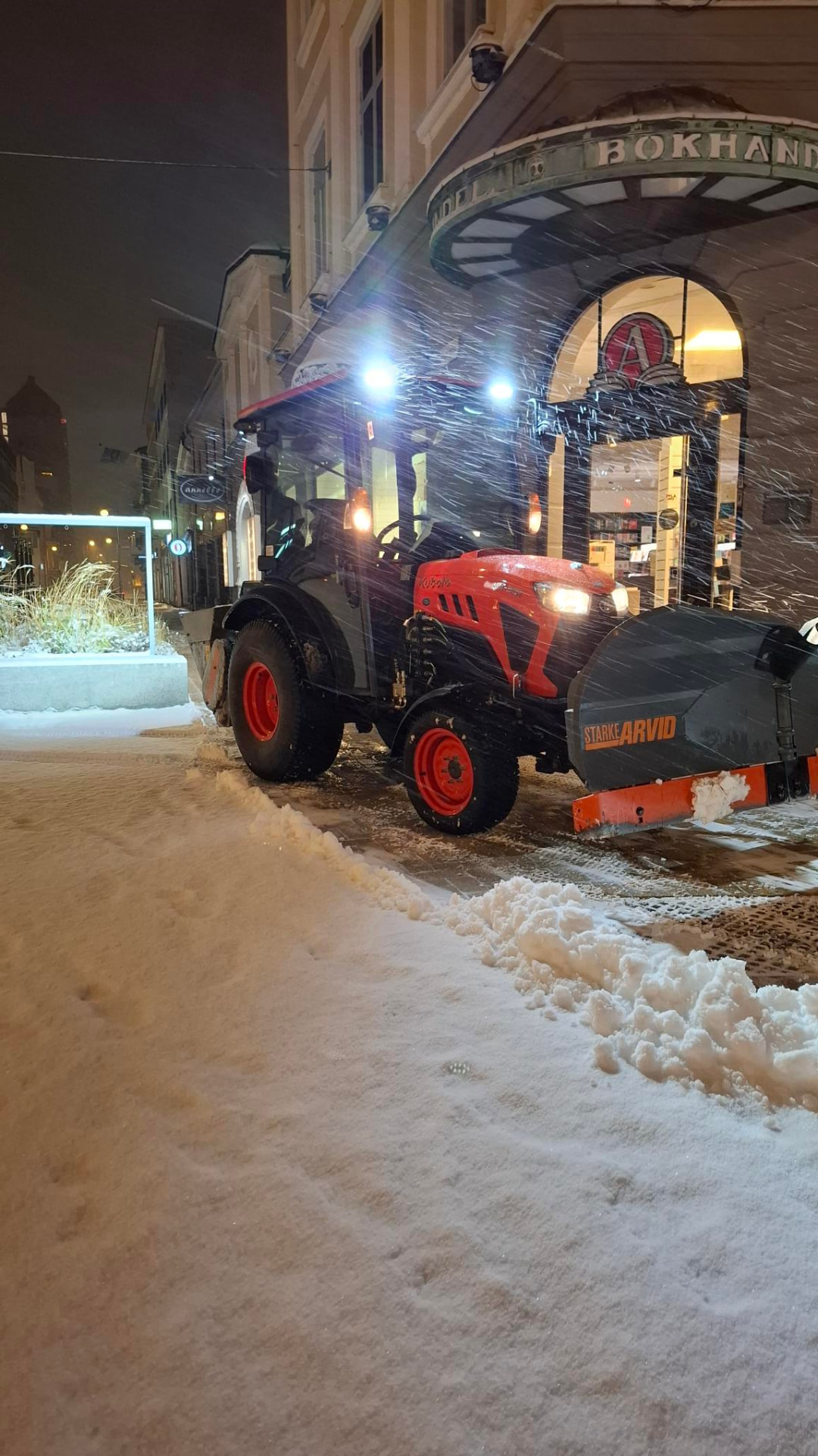 Traktor med snöplog röjer gångväg i snöfall kvällstid
